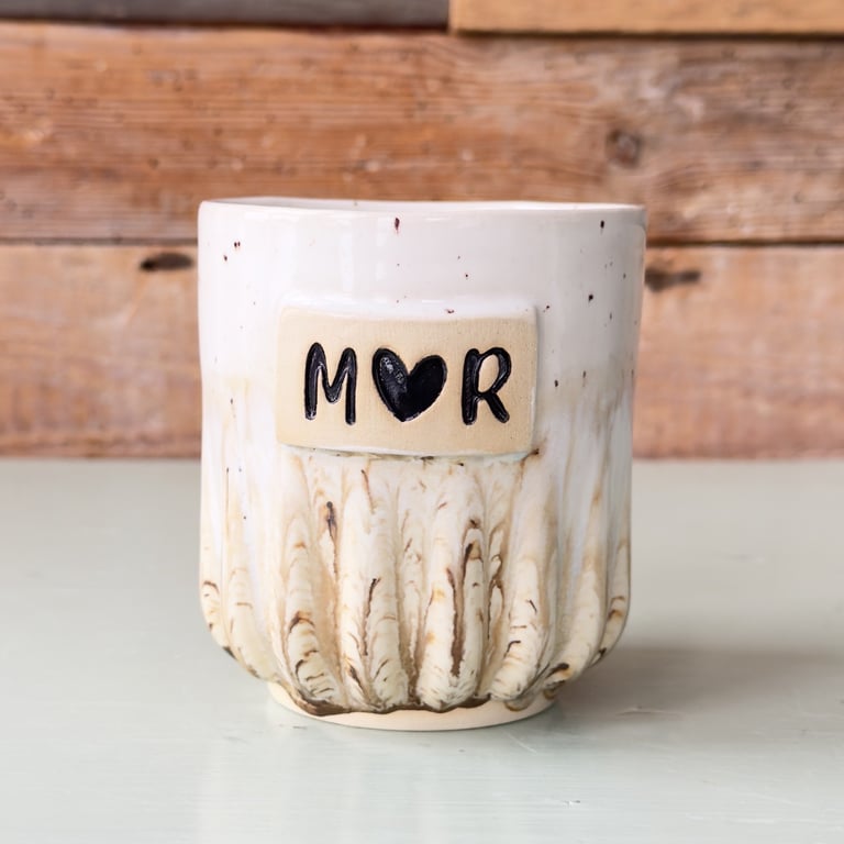 Keramikkrus med teksten "M♥R" på hvid og beige baggrund foran en trævæg.