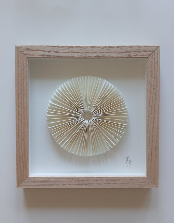Indrammet kunstværk med en rund, foldet papirdekoration i hvidt placeret på hvid baggrund, omgivet af en simpel træramme.