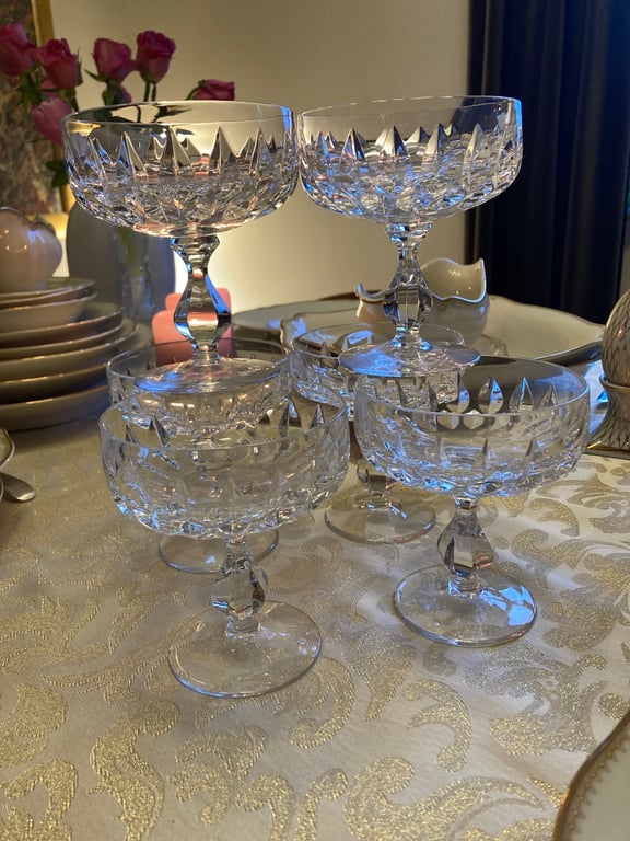 Fire elegante, slebne krystalglas i forskellig højde placeret på et dekoreret bord med tallerkener og blomster i baggrunden. (2)