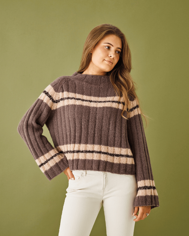 Kvinde iført en ribstrikket, brun trøje med beige og sorte striber, stående foran en grøn baggrund.