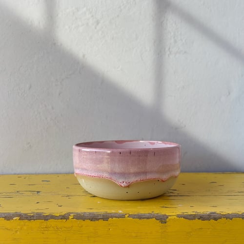 Lille keramisk skål med rosa og beige glasur placeret på et slidt gult træbord mod en hvid væg med skygger. (1)