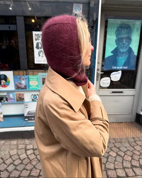 Kvinde med lyseblondt hår iført beige frakke og mørkerød strikhue, set fra siden, står på brostensbelagt gade foran en butiksvindue med plakater og bøger. (1)