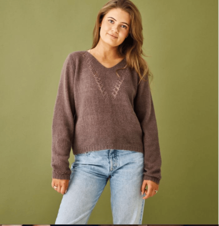 Ung kvinde iført brun strikket sweater og lyseblå jeans, stående foran grøn baggrund.