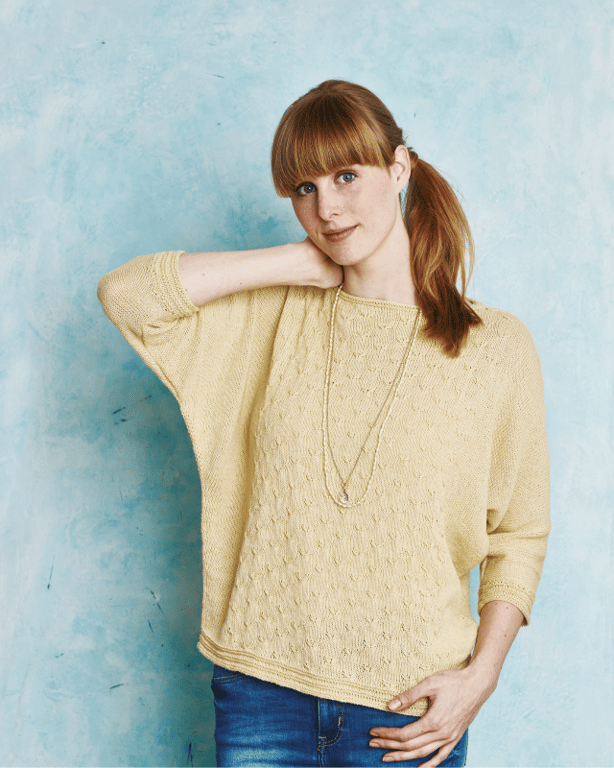 Portræt af en kvinde med rødt hår sat i en hestehale, iført en gul strikket trøje og blå jeans, stående foran en lyseblå væg.