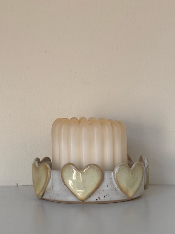 Et hvidt bloklys i en keramikstage med hjerteformer rundt om kanten, placeret på en hvid overflade mod en beige væg.