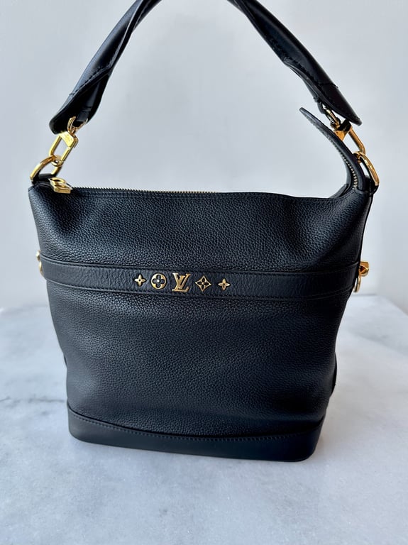 Sort Louis Vuitton håndtaske i læder med guld detaljer og kort hank, placeret på hvid og grå marmoroverflade.