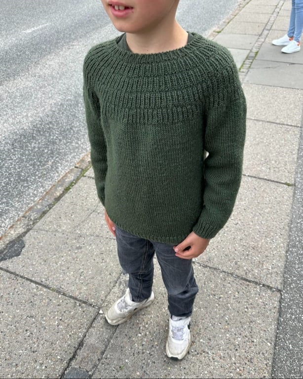 Dreng iført mørkegrøn striktrøje, mørke bukser og hvide sneakers står på fortov ved vej.