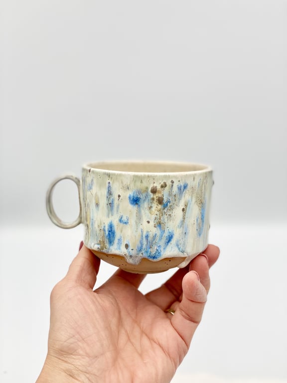 Hånd, der holder en håndlavet keramikkop med blå og brune pletter på en hvid baggrund.