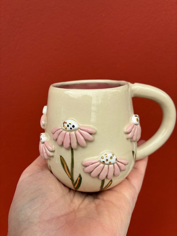 En cremefarvet keramik kop med lyserøde og hvide blomster dekorative elementer, holdt i hånd foran en rød baggrund.