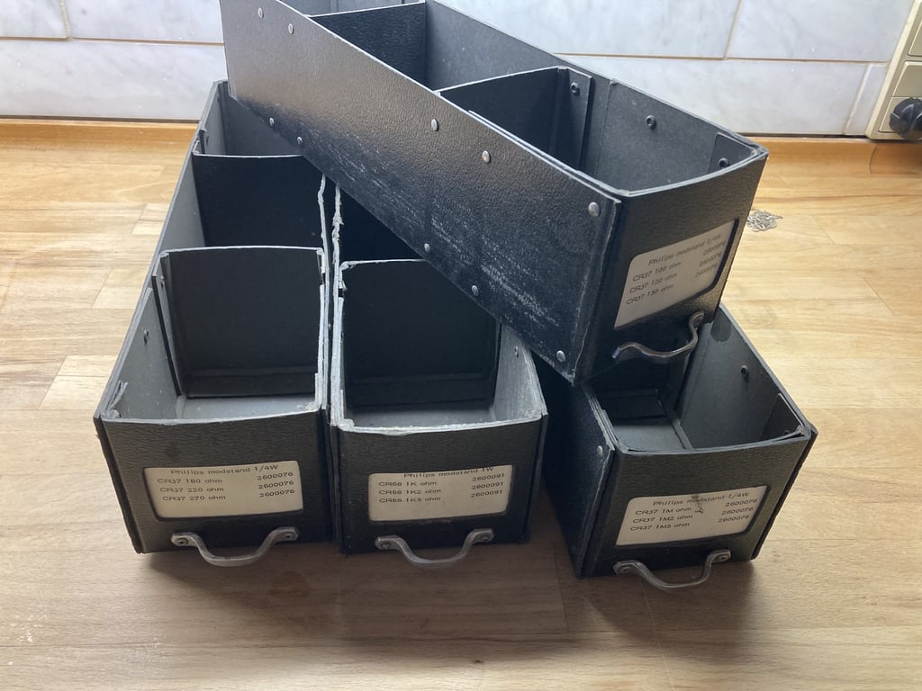 Fem sorte, firkantede metalbeholdere til opbevaring, arrangeret i en bunke på et lyst træbord. Labels med tekst i dansk er synlige på tre af beholderne. (2)