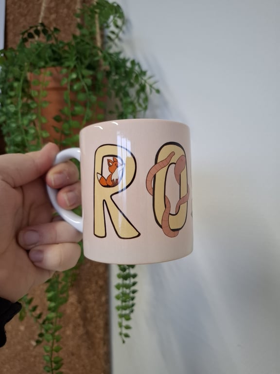 En hånd holder en lyserød krus med bogstaverne "RO" dekoreret med en rød ræv og en regnorm, med en grøn plante i baggrunden.