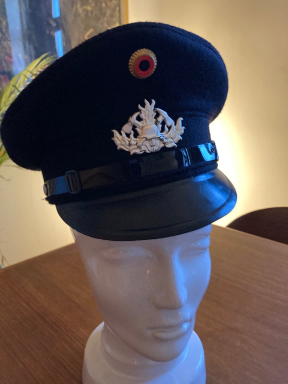 Sort uniformskasket med sølv emblem og rød, sort og gul rundel på en hvid mannequin hoved mod en baggrund med varmt lys. (2)