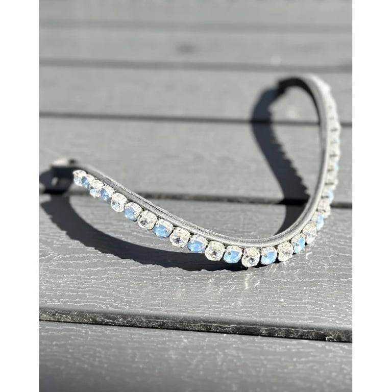 Billede af en hesteskoformet, sølvfarvet pandebånd med en række af klare og lyseblå perler, der ligger på en grå træoverflade i sollys.