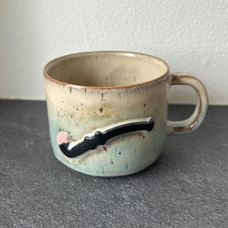 Keramisk kop med hank, beige og blålig glasur, dekoreret med relief af en sort pibe med rød glød.