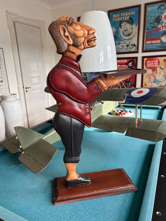 Træfigur af en mand med rød jakke og sorte bukser, der holder en bakke, placeret på et poolbord med en model af et militært fly og filmplakater i baggrunden.