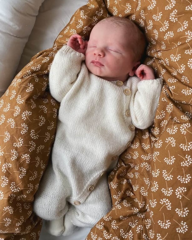 Nyfødt baby iført hvidt strikket heldragt, sovende på en brun dyne med hvidt blomstermønster.