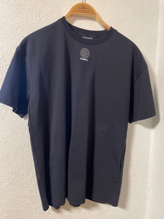 Sort t-shirt med rund hals og kortærmet, hængende på en træbøjle mod en hvid væg. Logo og teksten "Eskadron" er trykt foran på brystet.