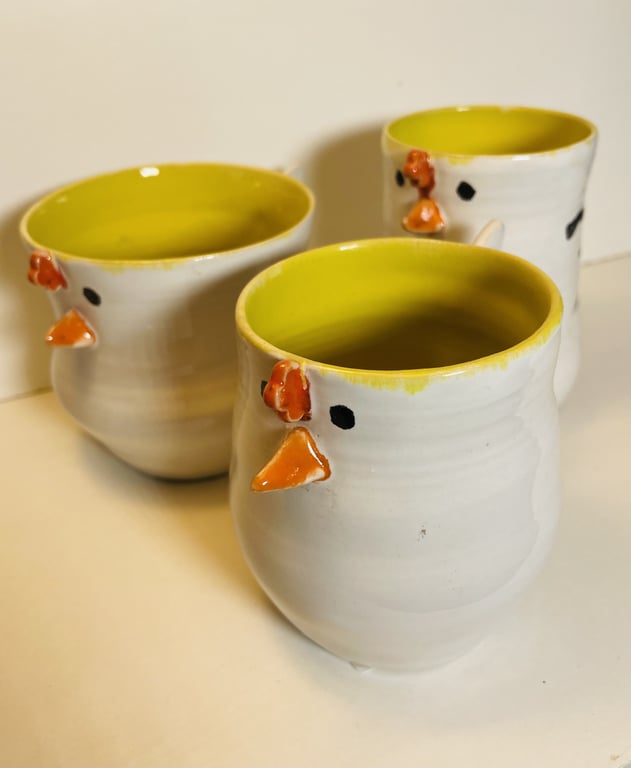 Tre hvide keramikkrus med gul inderside og dekoration, der ligner kyllingefacader med orangerødt næb og kam.