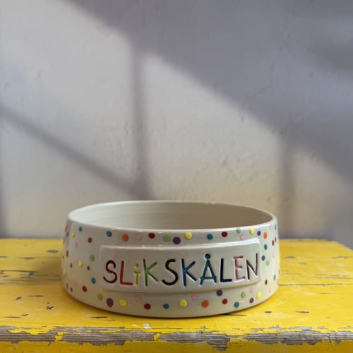 Keramikskål med farverige prikker og teksten "SLIKSKÅLEN" i forskellige farver, placeret på et slidt, gult træbord. (1)