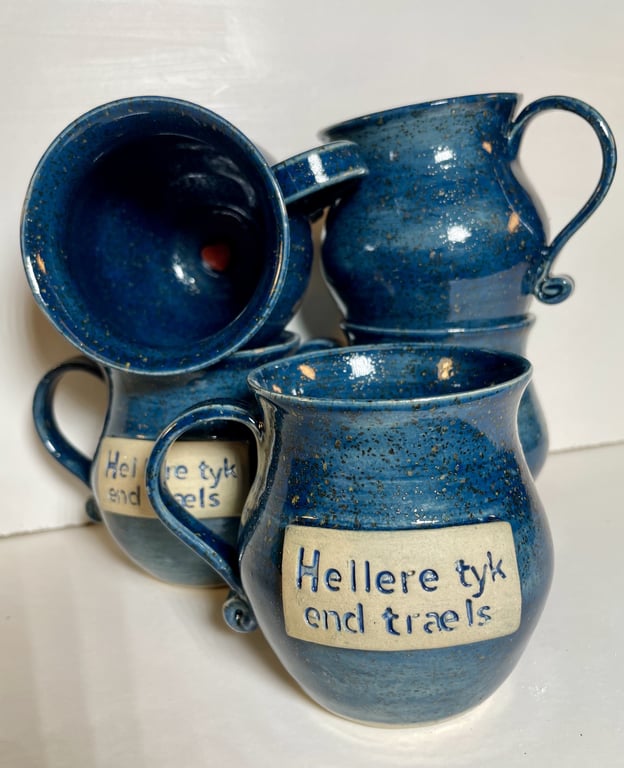 Fire blå keramikkrus med hank og teksten "Hellere tyk end træls" på en beige plade på krusene.