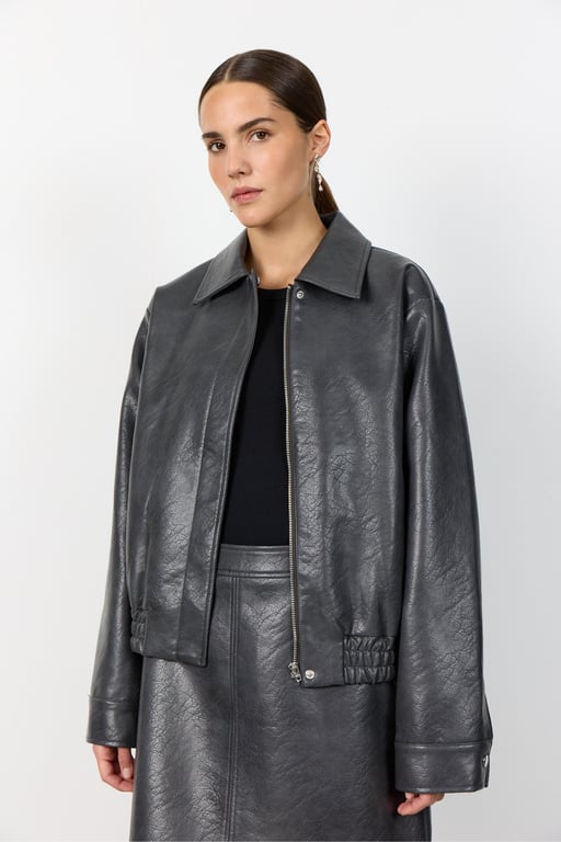 Kvinde iført sort læderjakke og nederdel med sort top, stående foran hvid baggrund.