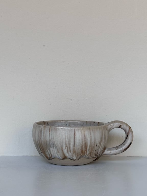 Keramisk kop med rund hank og marmoreret gråbrun glasur, placeret på hvid overflade med hvid væg i baggrunden.