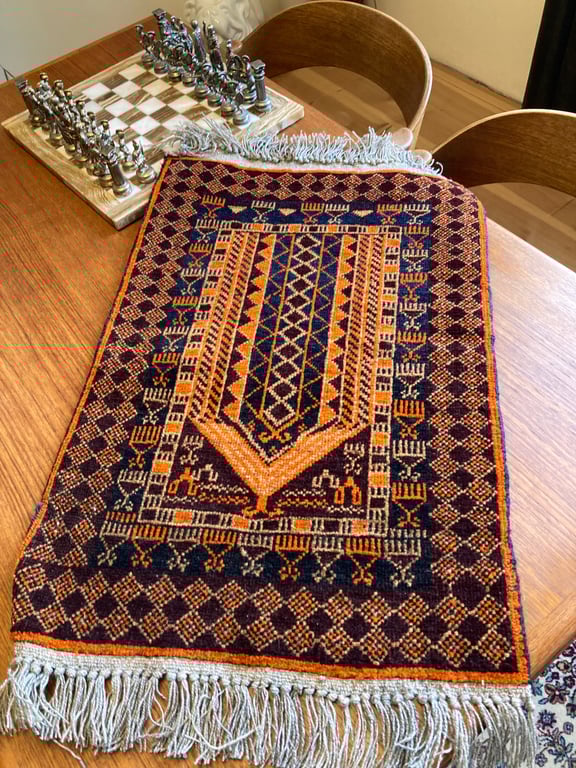 Farverigt, mønstret tæppe med frynser på et træbord ved siden af et metal skakspil og to træstole. (2)