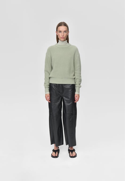 Kvinde iført lysegrøn ribstrikket sweater, sorte læderbukser og sorte sandaler, stående mod en grå baggrund.