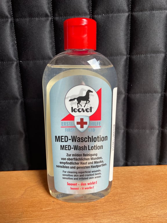 Flaske med Leovet MED-Wash Lotion til mild rengøring af overfladiske sår og sensitiv hud, står på et træbord med en sort quiltet baggrund.