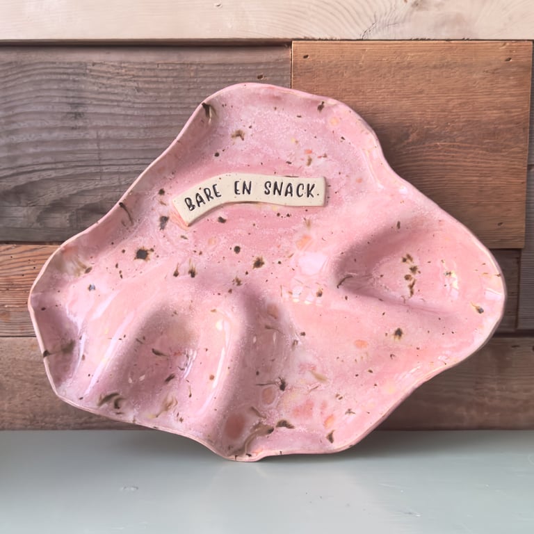 Pink, uregelmæssigt formet keramikfad med teksten "BÅRE EN SNACK" påsat, placeret foran en trævæg.