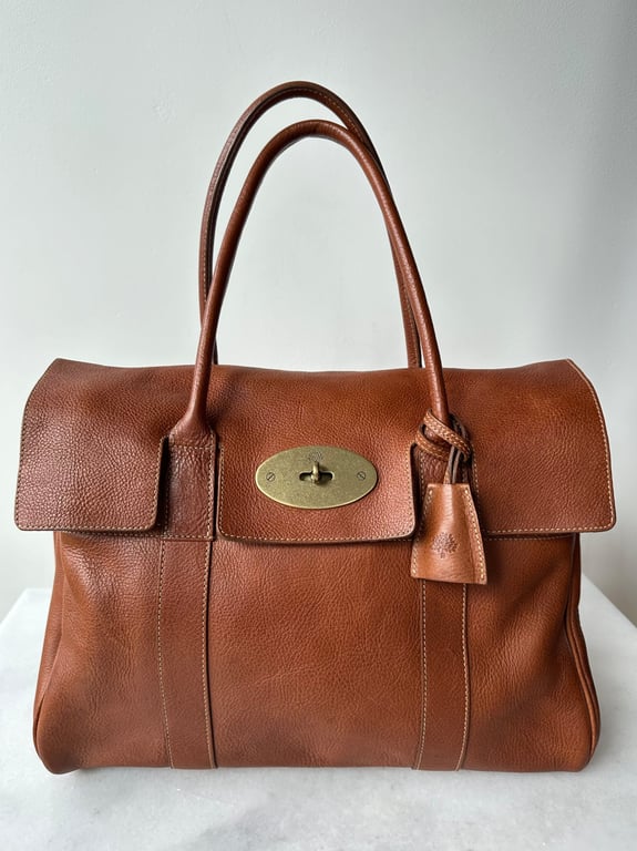 Brun lædertaske med to håndtag og metalspænde foran på hvid baggrund.