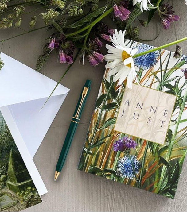Billede af en bog med titel "Anne Just" med blomster på forsiden, en grøn og gul pen, et åbent hvidt kuvert og blomster, herunder en marguerit, lagt på et bord.