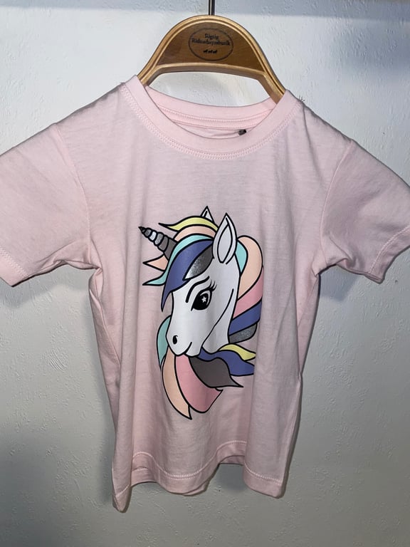 Lyserød børne T-shirt med illustration af enhjørning med pastelfarvet manke og store øjne, hængende på en træbøjle mod hvid væg.