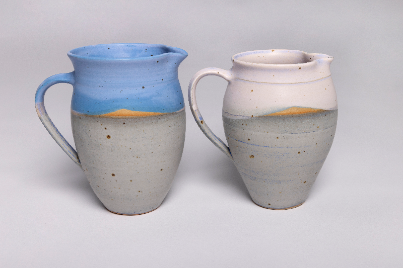 To keramiske krus med håndtag, den venstre er blå med en beige bund og en blå himmel på toppen, den højre er hvid med en beige bund og en lys himmel på toppen. (3)