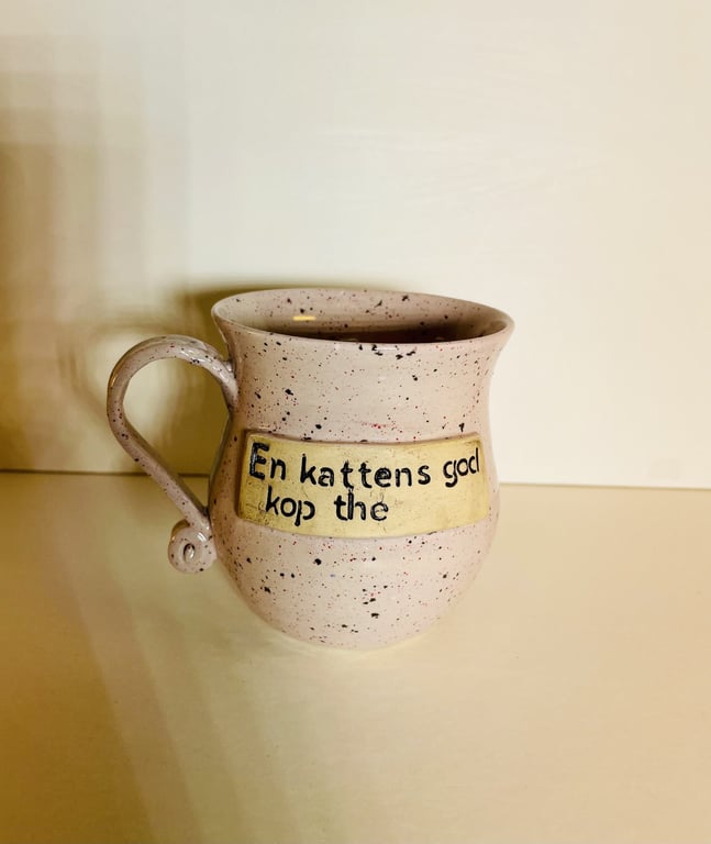 Keramikkrus med tekst: "En kattens god kop the", på beige overflade med sorte prikker.