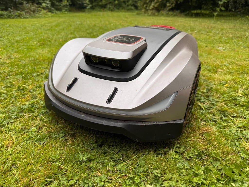 Robotplæneklipper på græsplæne med buske og træer i baggrunden.