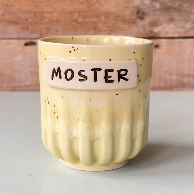 Gul keramikkop med teksten "MOSTER" på, placeret på en lys overflade med en træbaggrund.