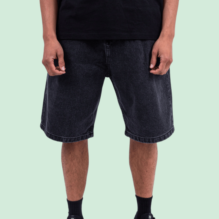 Mand iført sorte jeansshorts og sort t-shirt stående mod en lysegrøn baggrund, kun synlig fra skuldre til fødder.