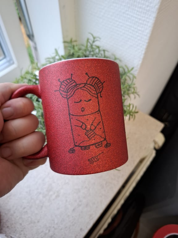 Rød glitrende kop med sort tegning af en sød, sovende figur med to garnnøgle-knuder som hår.