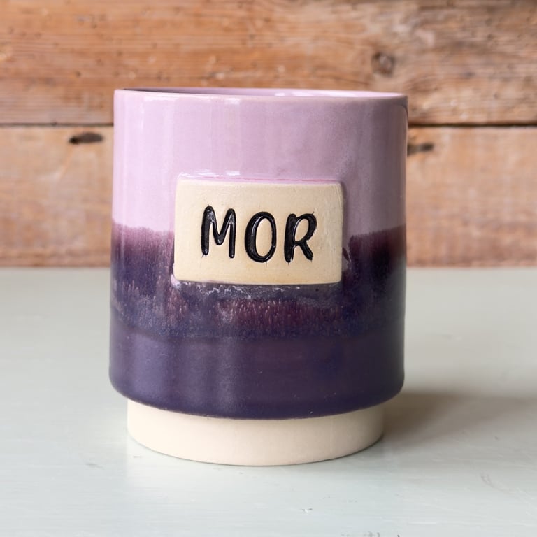 Keramikkrus med farver i lilla nuancer og teksten "MOR" på en beige firkant i midten.