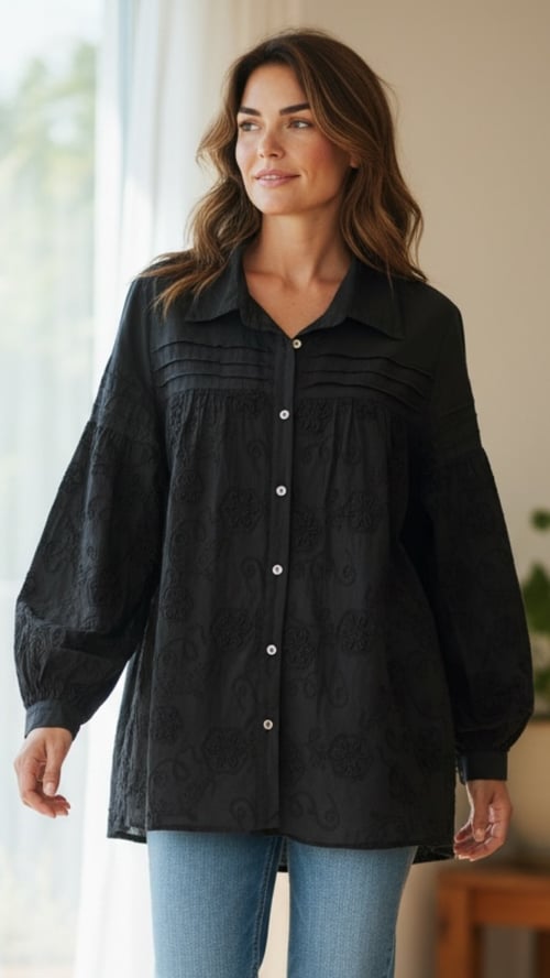 Kvinde iført sort langærmet knapbluse med broderede detaljer og blå jeans, stående ved lyse gardiner. (1)