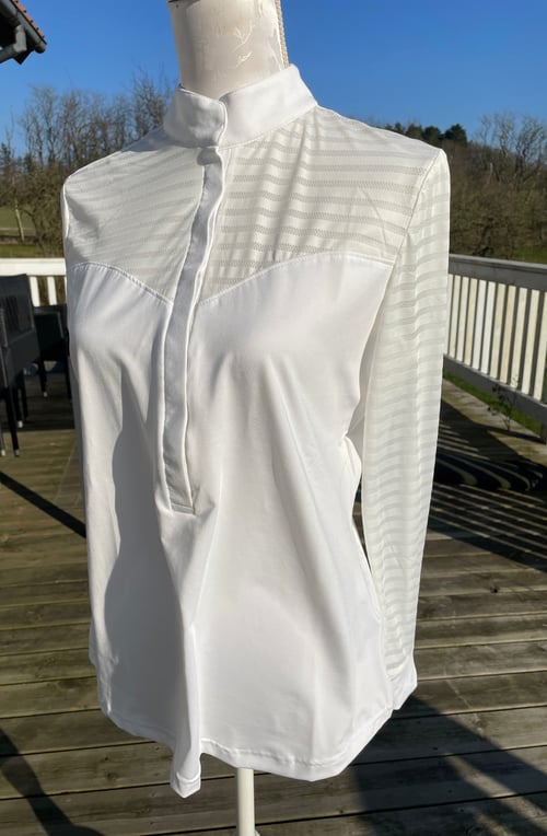 Hvid langærmet skjorte med mønsterdetaljer på skuldre og ærmer, afsluttet med en kort stående krave, vist på en mannequin udendørs på en træterrasse. (1)