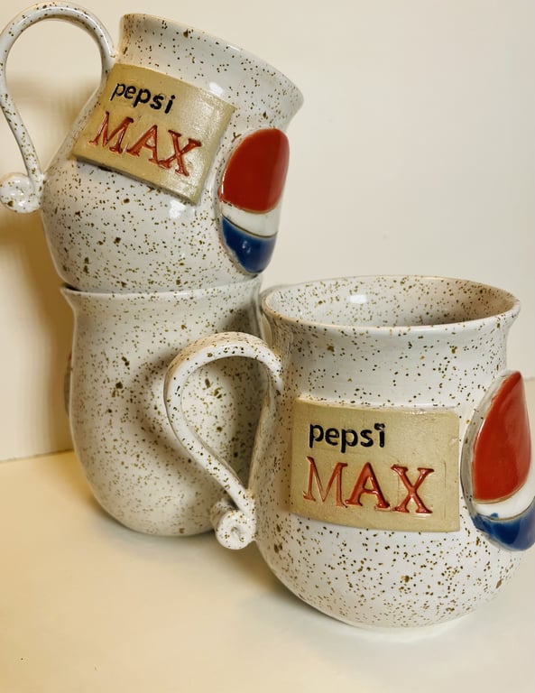 To keramiske kaffe- eller tekopper med "Pepsi Max" logo og plettet, beige overflade.