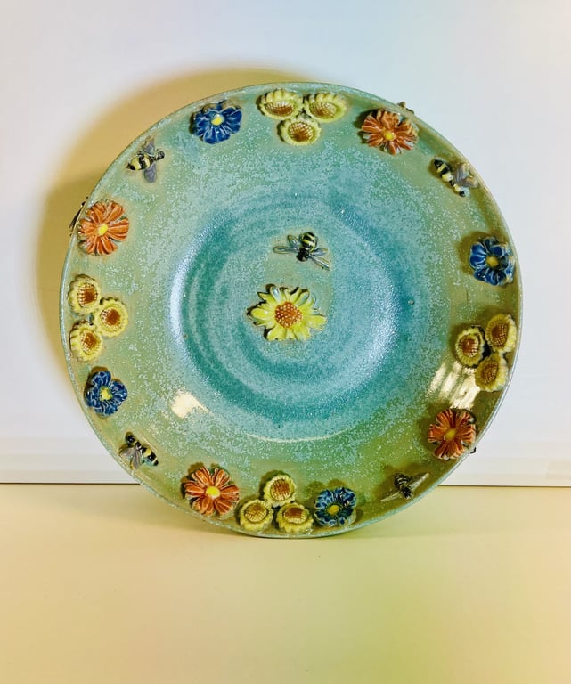 Rundt, grønt keramiktallerken med reliefdekoration af farverige blomster og insekter.