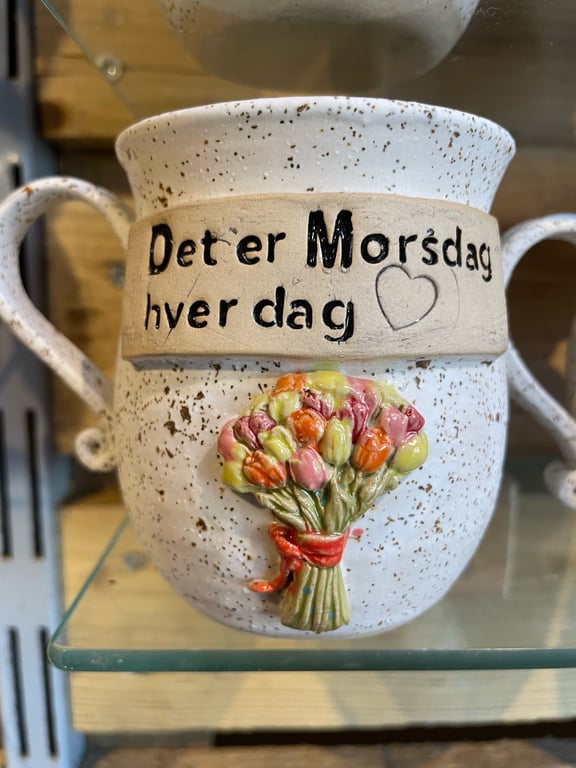 Hvidt, keramisk krus med brun tekstur med teksten "Det er Morsdag hver dag" og et relief af en farverig buket blomster bundet med et rødt bånd.