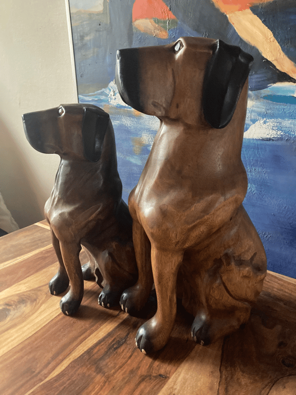 To træhundefigurer med mørke ører, siddende på et træbord foran et maleri. (2)