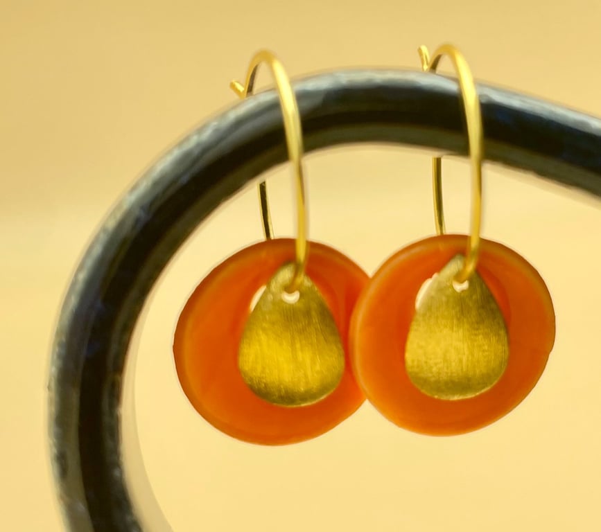 Nærbillede af et par øreringe med gule metaldråber og orange cirkler, hængende på en sort bøjle.