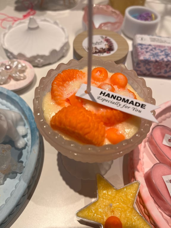 Lys dekorativ dessert i en glas-skål med orange frugt og et mærkat med teksten "HANDMADE Especially for You", omgivet af andre små dekorationer og stearinlys.
