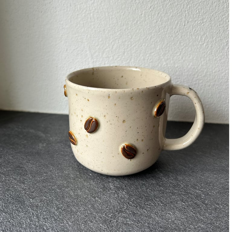 Keramisk beige kaffekop med håndtag og dekorative kaffebønner på ydersiden, placeret på mørk bordplade foran hvid væg.