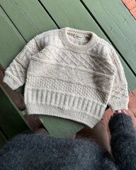 En lys beige, strikket sweater med mønster, lagt ud på en grøn trægulv. En person i en mørk trøje holder i kanten af sweateren. (1)